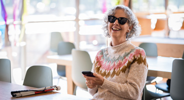 A woman smiles as she listens to NFB-NEWSLINE on her phone in a cafe.